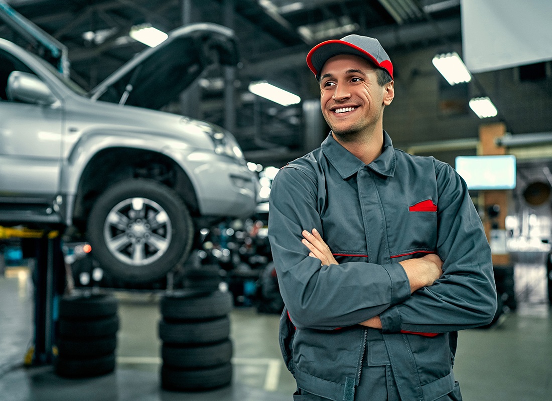 Insurance by Industry - Portrait of a Cheerful Young Mechanic Standing in a Garage Repair Shop with his Arms Folded in Front of a Car up on a Lift