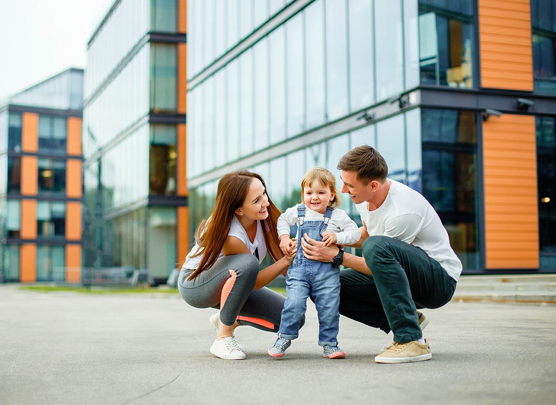 Personal Insurance - Portrait of Two Young Parents Having Fun Holding Their Toddler as They Stand Outside a Modern Building in the City